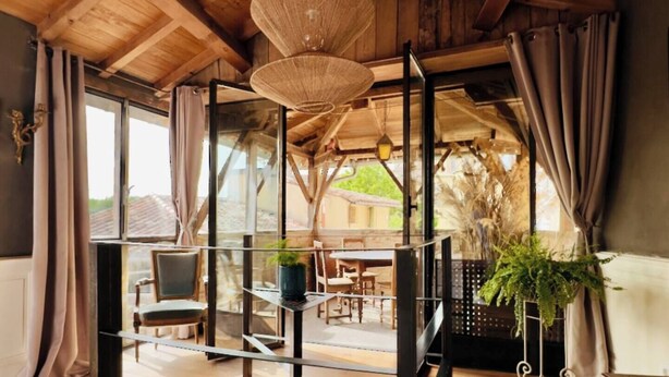 Une pièce avec un plafond en bois, de grandes fenêtres et une vue sur l’extérieur. On y trouve des chaises et une table sur laquelle est posée une plante en pot.
