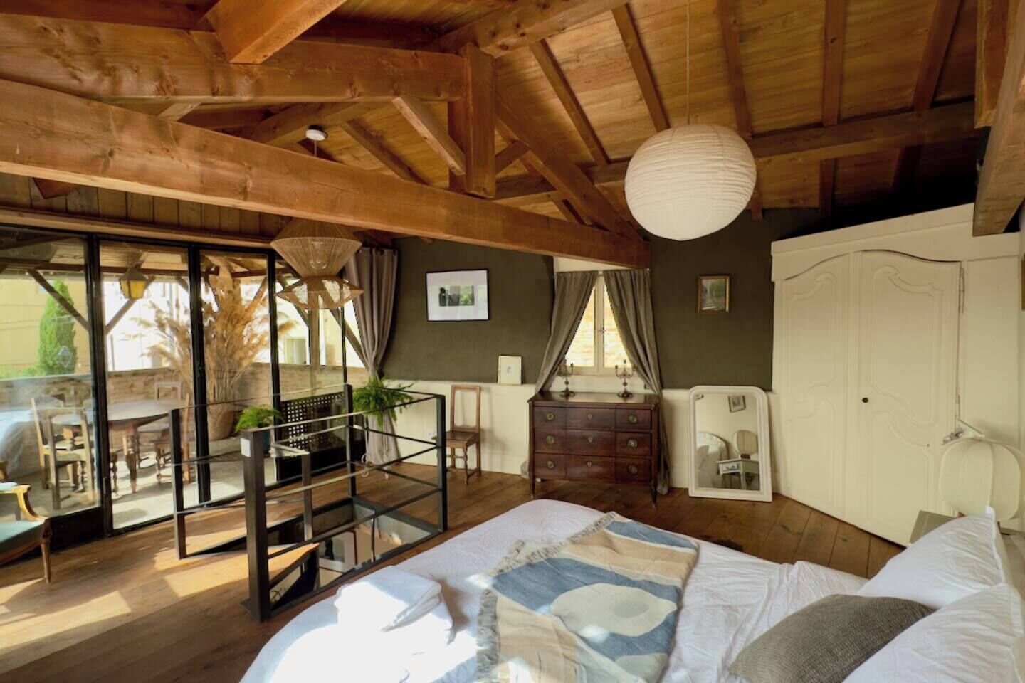 A bedroom with a wooden ceiling, a bed with a patterned bedspread, a dresser, a mirror, and a window with curtains.