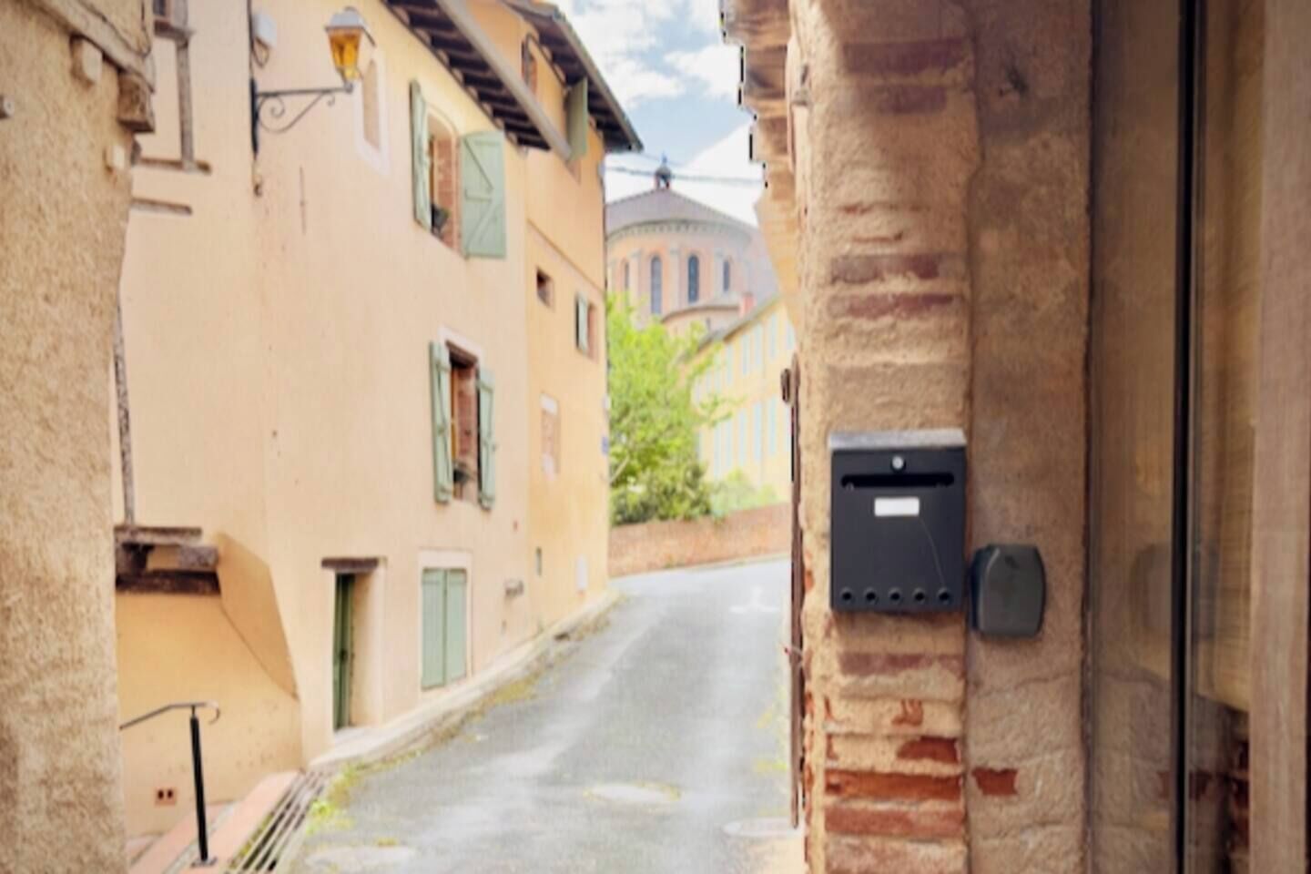 A narrow street with buildings on either side, a mailbox attached to a brick wall, and a dome-shaped building in the background.