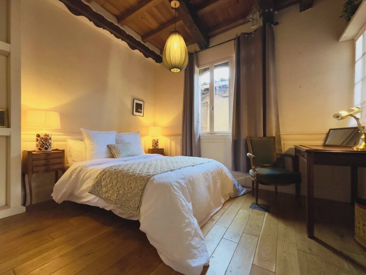 Un dormitorio con piso de madera, una cama con ropa de cama blanca, un escritorio con silla y una ventana con cortinas.