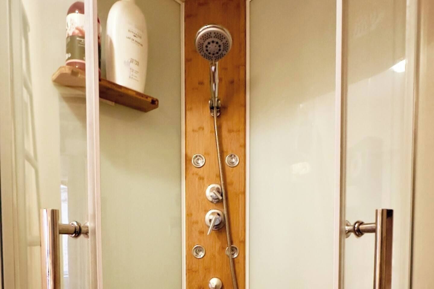 A shower with a wooden panel, a handheld showerhead, and a bottle of shampoo on a shelf.