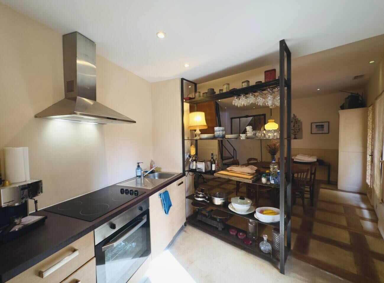 Een compacte keuken met een fornuis, oven en een moderne afzuigkap. Achtergrond zichtbaar: een eethoek met tafel en stoelen.