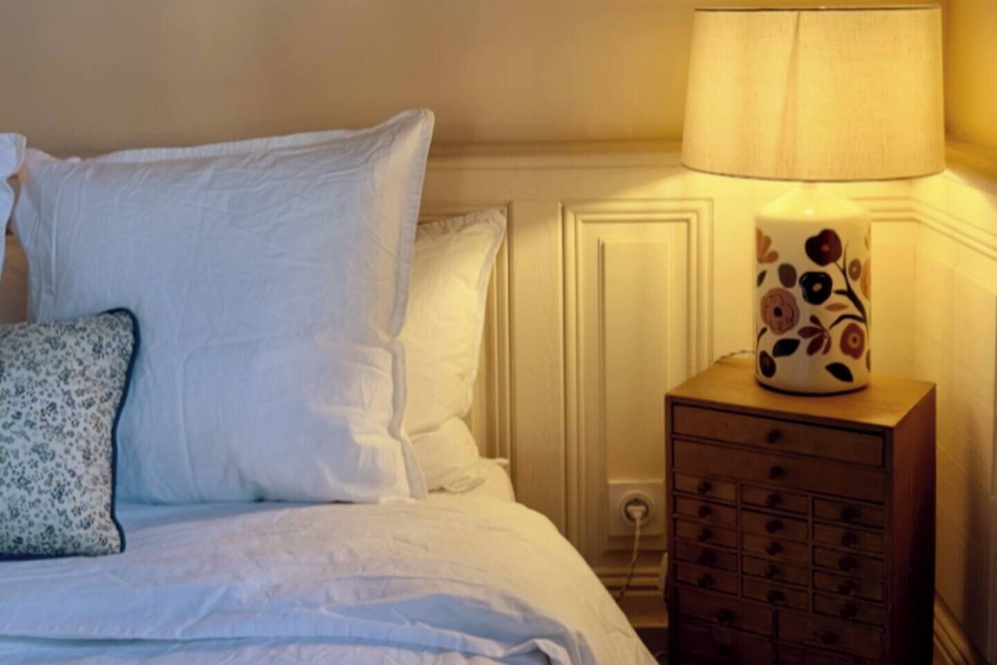 Un letto rifatto con lenzuola bianche e una lampada a motivi floreali su un comodino in legno.