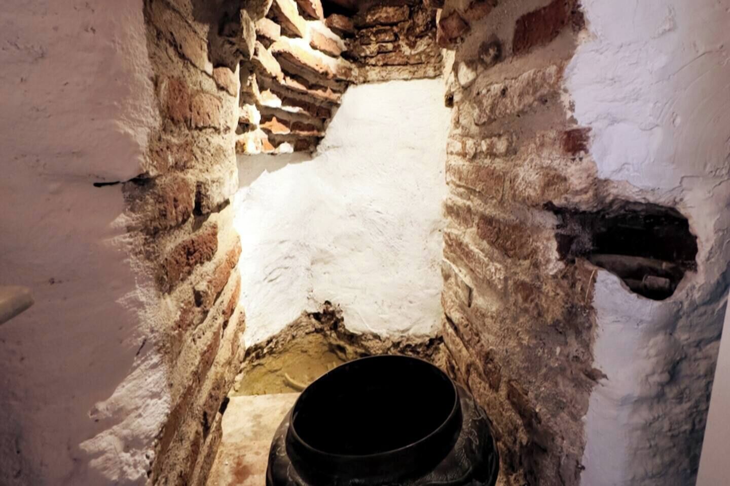 Een smalle, stenen doorgang met een ruw, ongelukkig plafond en een grote, donkere pot onderin.