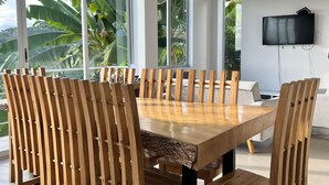 Ein moderner Essbereich mit einem großen Holztisch und Stühlen, großen Fenstern mit Blick auf Grünflächen und einem an der Wand montierten Flachbildfernseher.
