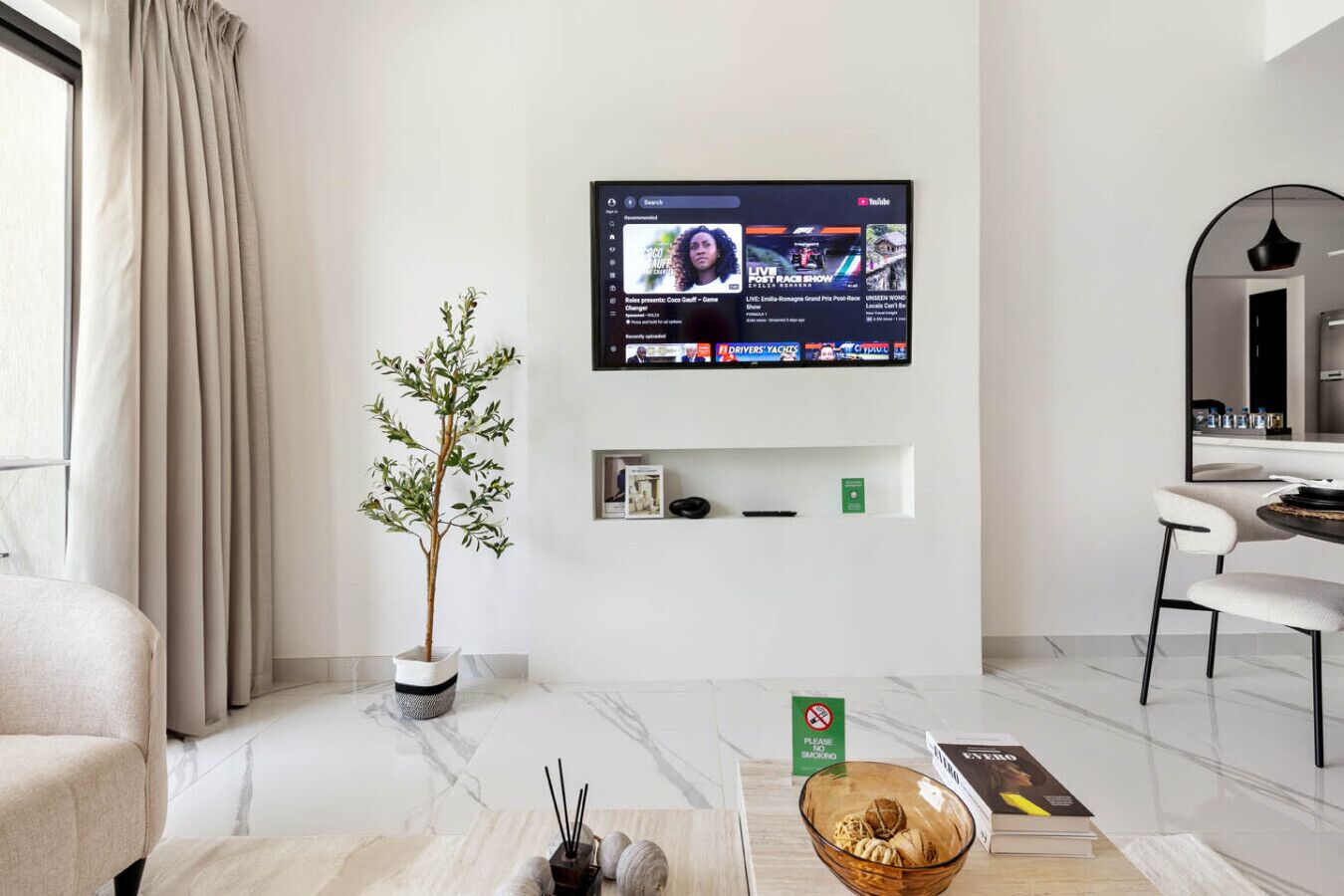 A modern living room with a flat-screen TV mounted on the wall, a potted plant, a small shelf with books, a bowl of snacks, and a dining area with a chair and table.