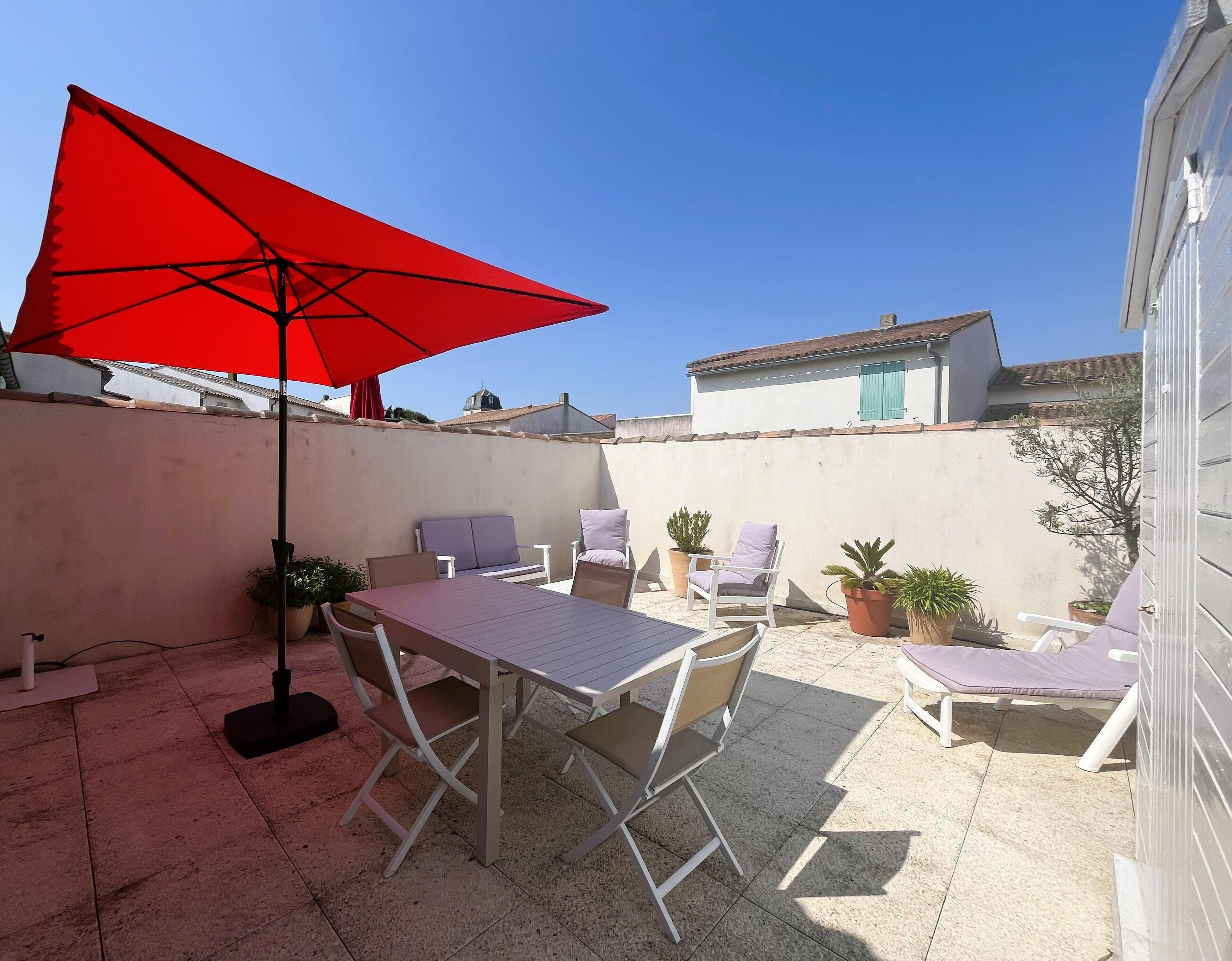 A patio with a table and chairs, a red umbrella, and potted plants.