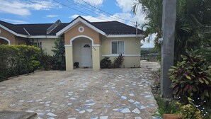 Una casa con un camino de entrada empedrado que conduce a una puerta blanca, rodeada de follaje y un cielo despejado.