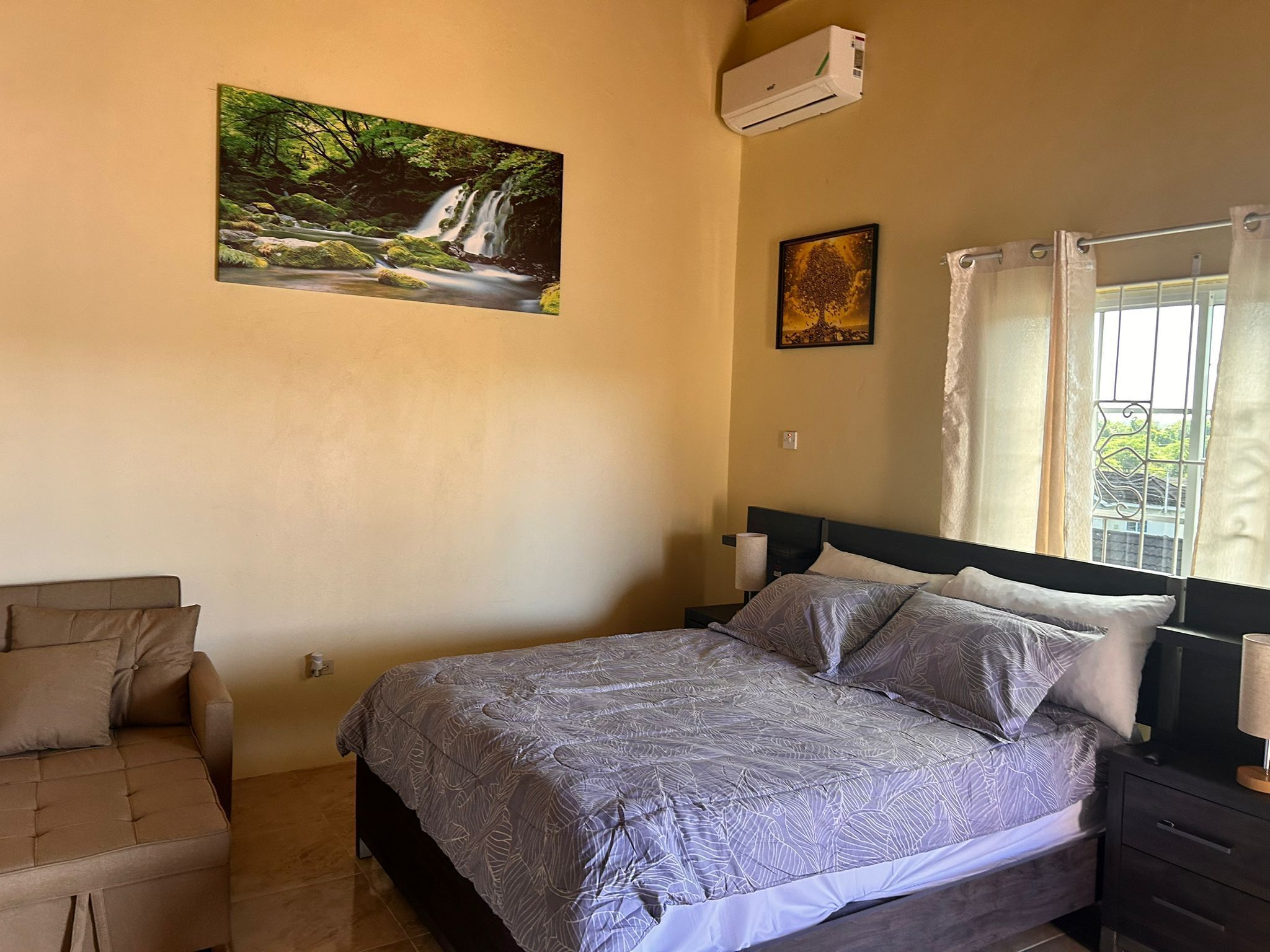 A bedroom with a bed, a chair, a painting of a waterfall, and a window with curtains.