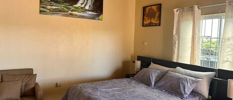 A bedroom with a bed, a chair, a painting of a waterfall, and a window with curtains.