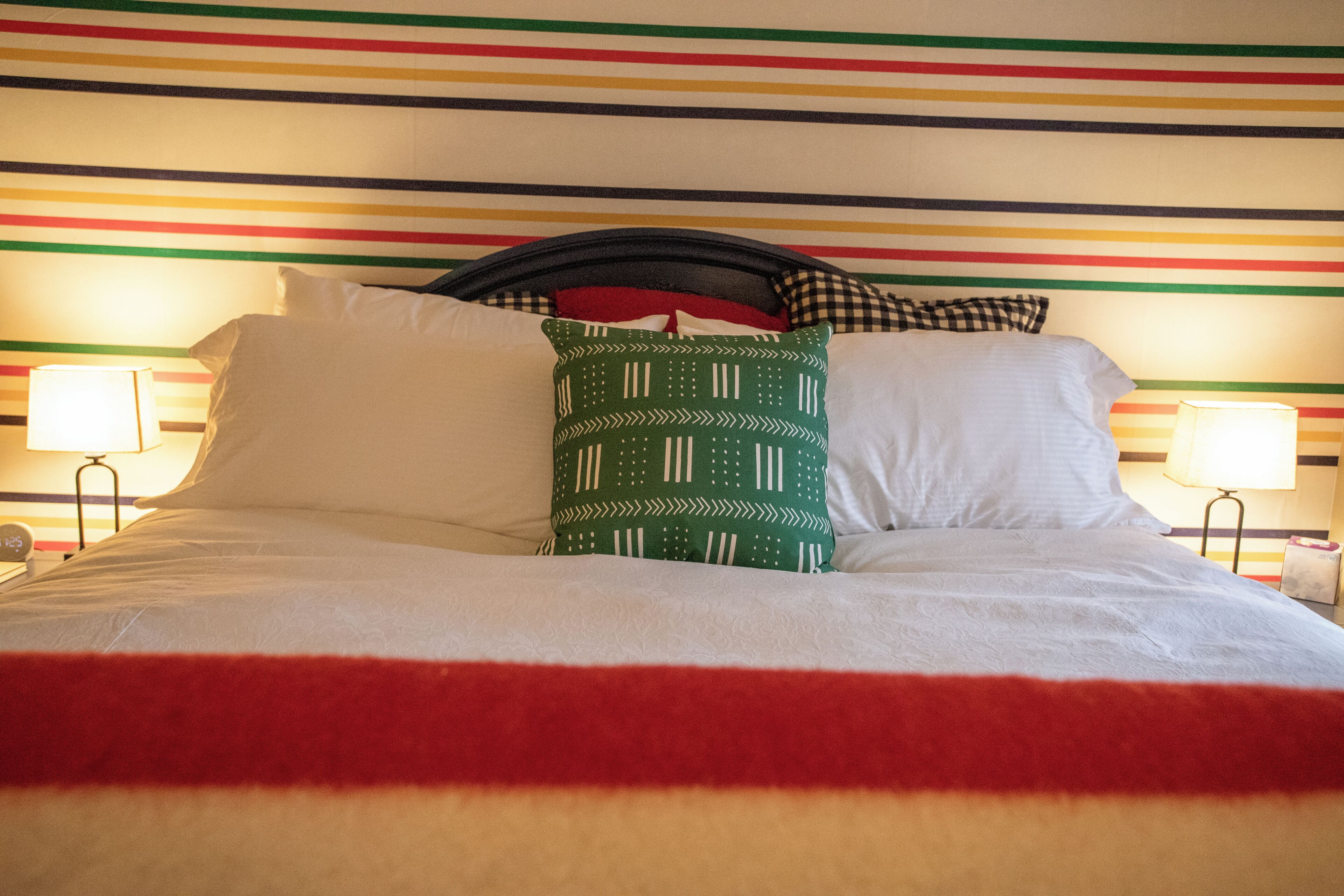 A bed with white bedding and a red blanket, a green patterned pillow, and a black headboard.