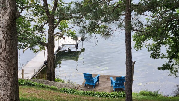 A dock extending into a calm lake, surrounded by trees. Two blue chairs are placed on a wooden platform near the water's edge.