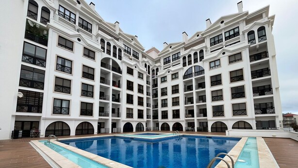 Uma piscina grande e retangular com um deck ladrilhado ao redor, com um edifício de vários andares e varandas e janelas em arco ao fundo.