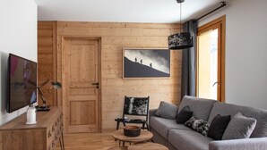A cozy living room with a grey sofa, wooden coffee table, and a black chair. A wooden door and a framed picture of hikers are also visible.