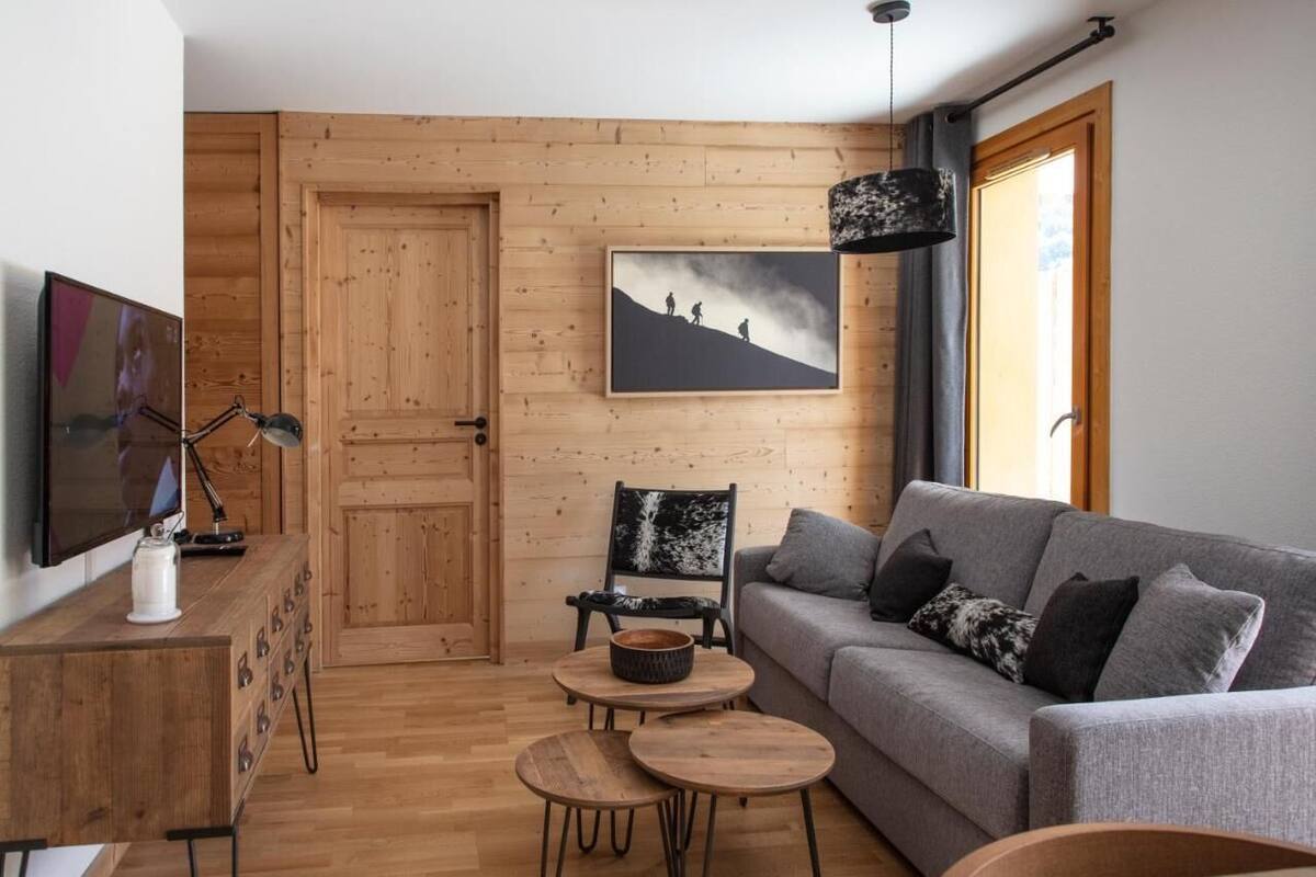 Ein gemütliches Wohnzimmer mit grauem Sofa, Holztisch und schwarzem Sessel. Eine Holztür und ein gerahmtes Bild von Wanderern sind ebenfalls zu sehen.