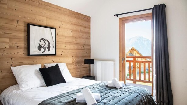 A bedroom with a wooden headboard, a framed picture, a bed with white and black bedding, a grey quilt, and a view of a wooden deck and mountains through a window.