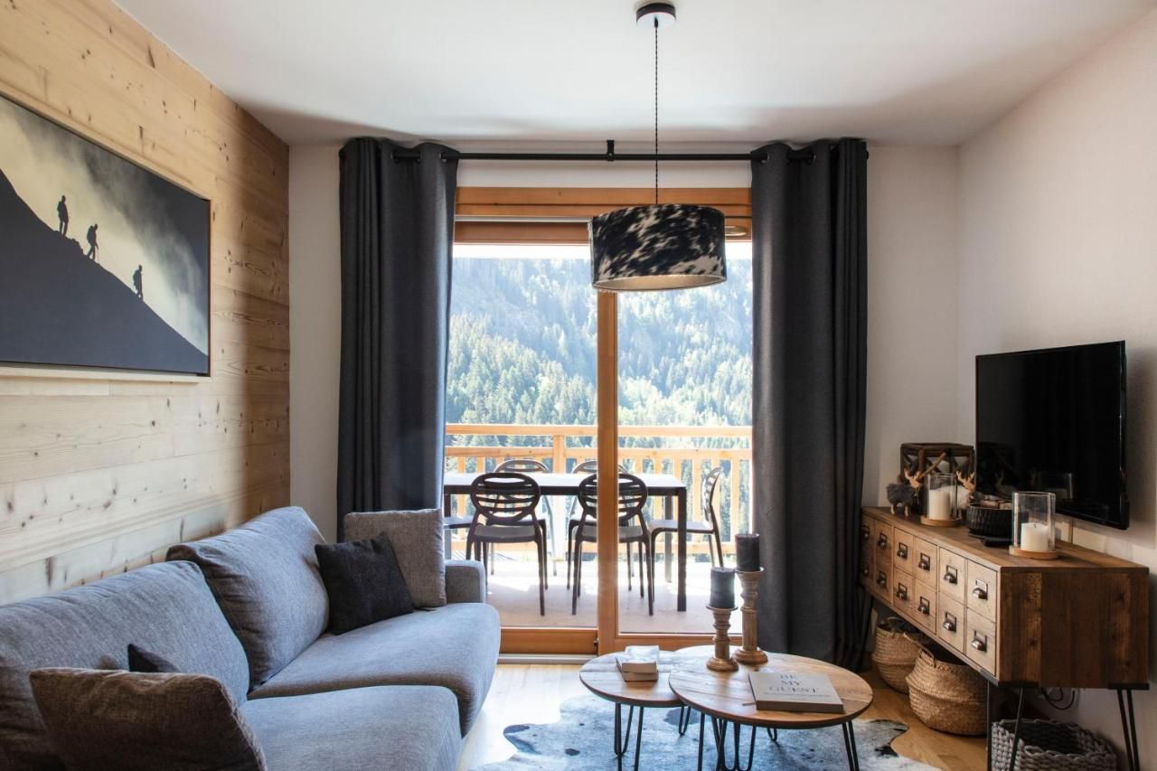 Un salon moderne comprenant un canapé, une table à manger avec des chaises, un téléviseur et une vue sur une montagne boisée, visible à travers une grande fenêtre.