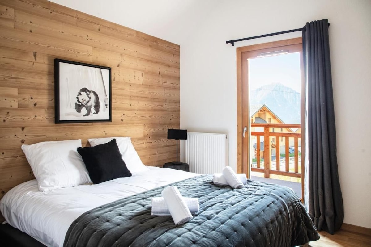 Un dormitorio con cabecera de madera, un cuadro enmarcado, ropa de cama blanca y negra, una colcha gris y vistas a una terraza de madera y montañas a través de una ventana.