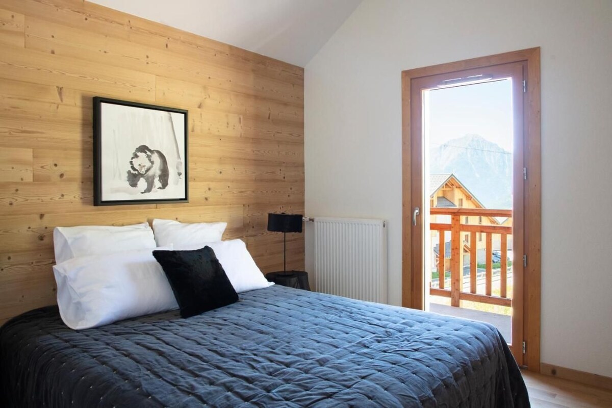 A bedroom with a wooden headboard, a framed picture, a bed with white and black pillows, a bedside table with a lamp, and a view of a wooden balcony with mountains in the background.