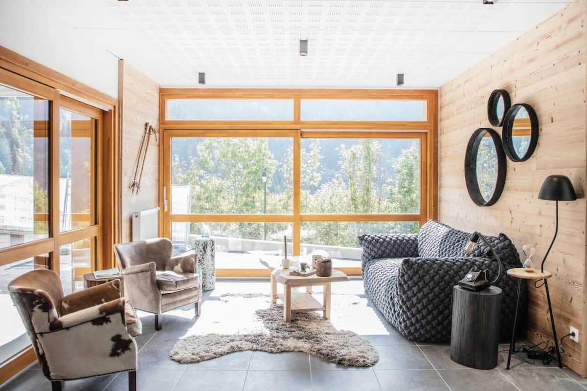 Ein modernes Wohnzimmer mit großen Fenstern, einer Couch, Sesseln und einem Couchtisch.