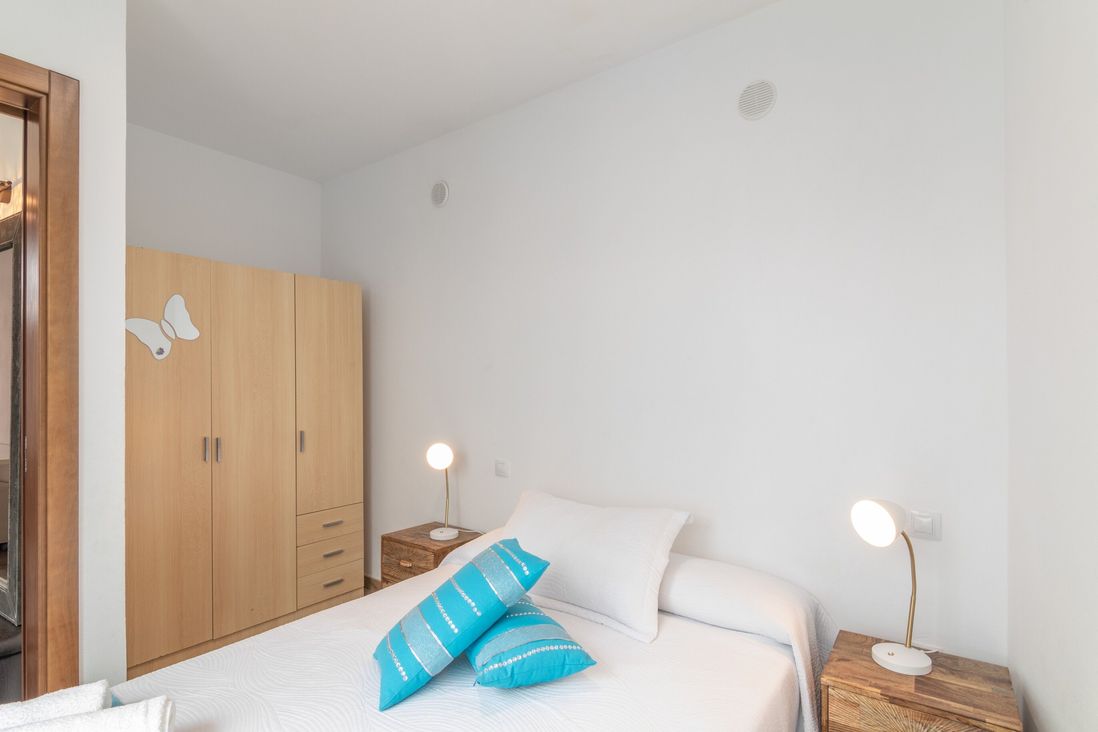 Un dormitorio con cama, dos mesitas de noche con lámparas, un armario y una pared con un respiradero.