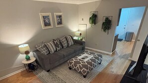 Un salón con un sofá gris, un reposapiés estampado y una mesita auxiliar con una lámpara. Se observan dos cuadros en la pared, una lámpara de pie y una planta en maceta.