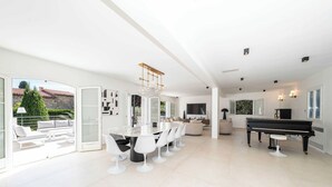 A spacious living area with a piano, a dining table with chairs, and a large window overlooking a garden.