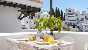 Une table blanche avec des chaises sur une terrasse, un petit arbre et, en arrière-plan, une vue sur des bâtiments blancs.
