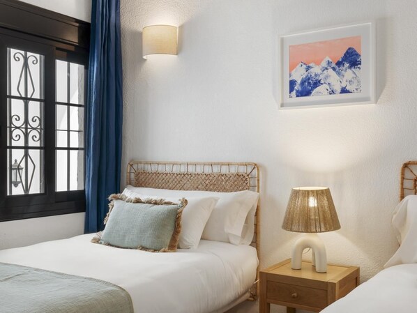 A bedroom with a bed, a nightstand, a wall-mounted lamp, a window with curtains, and a framed picture on the wall.
