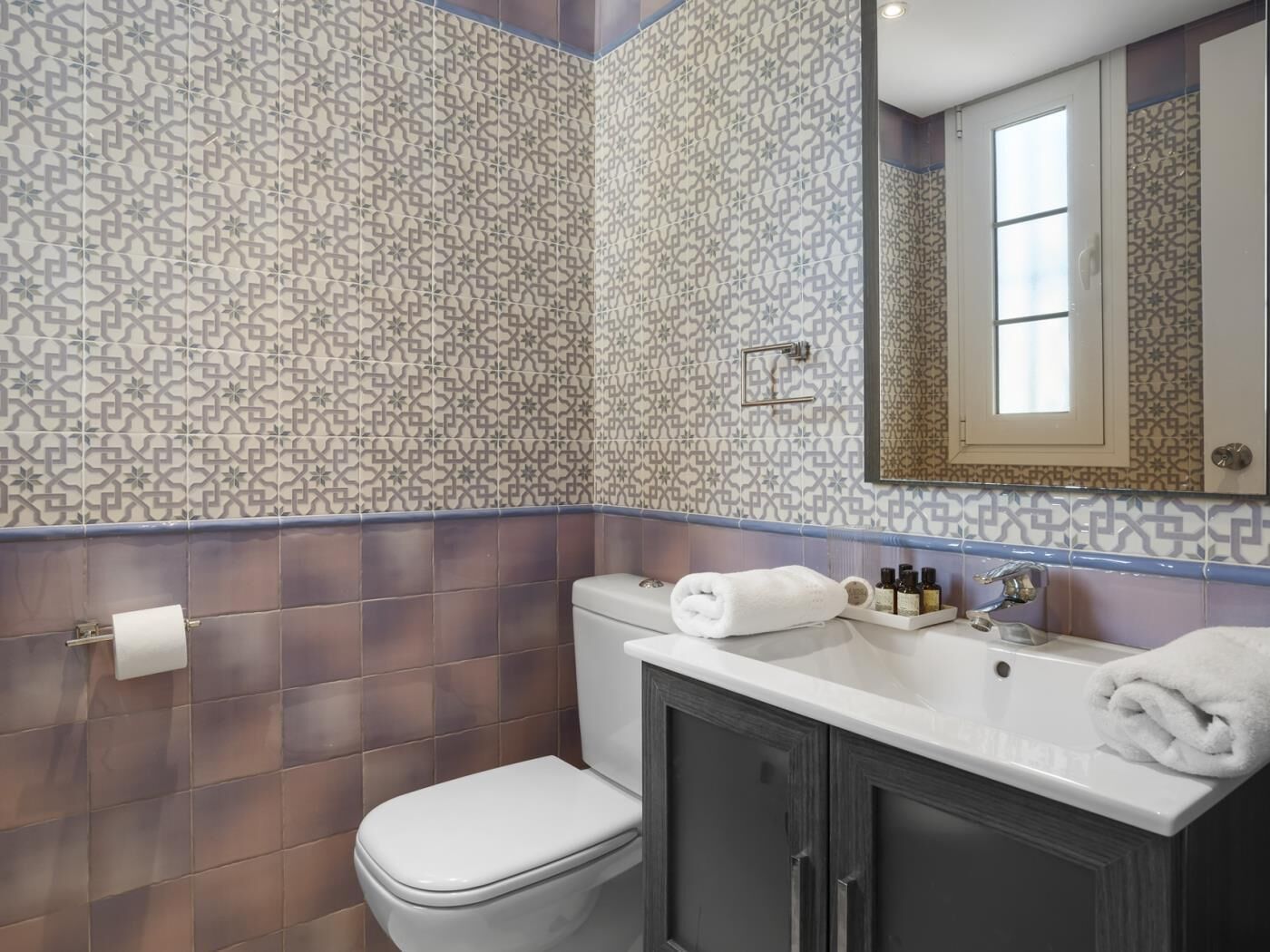 A bathroom with patterned tiles, a toilet, a sink with toiletries, and a mirror.