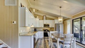 Una cocina moderna con armarios blancos, encimera de granito y electrodomésticos de acero inoxidable. Cuenta con un comedor con mesa de madera y sillas blancas.