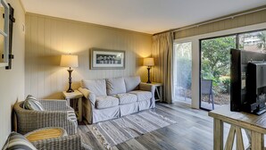 Un salón acogedor con un sofá, dos mesitas de noche con lámparas, un sillón de mimbre, una alfombra y un televisor.