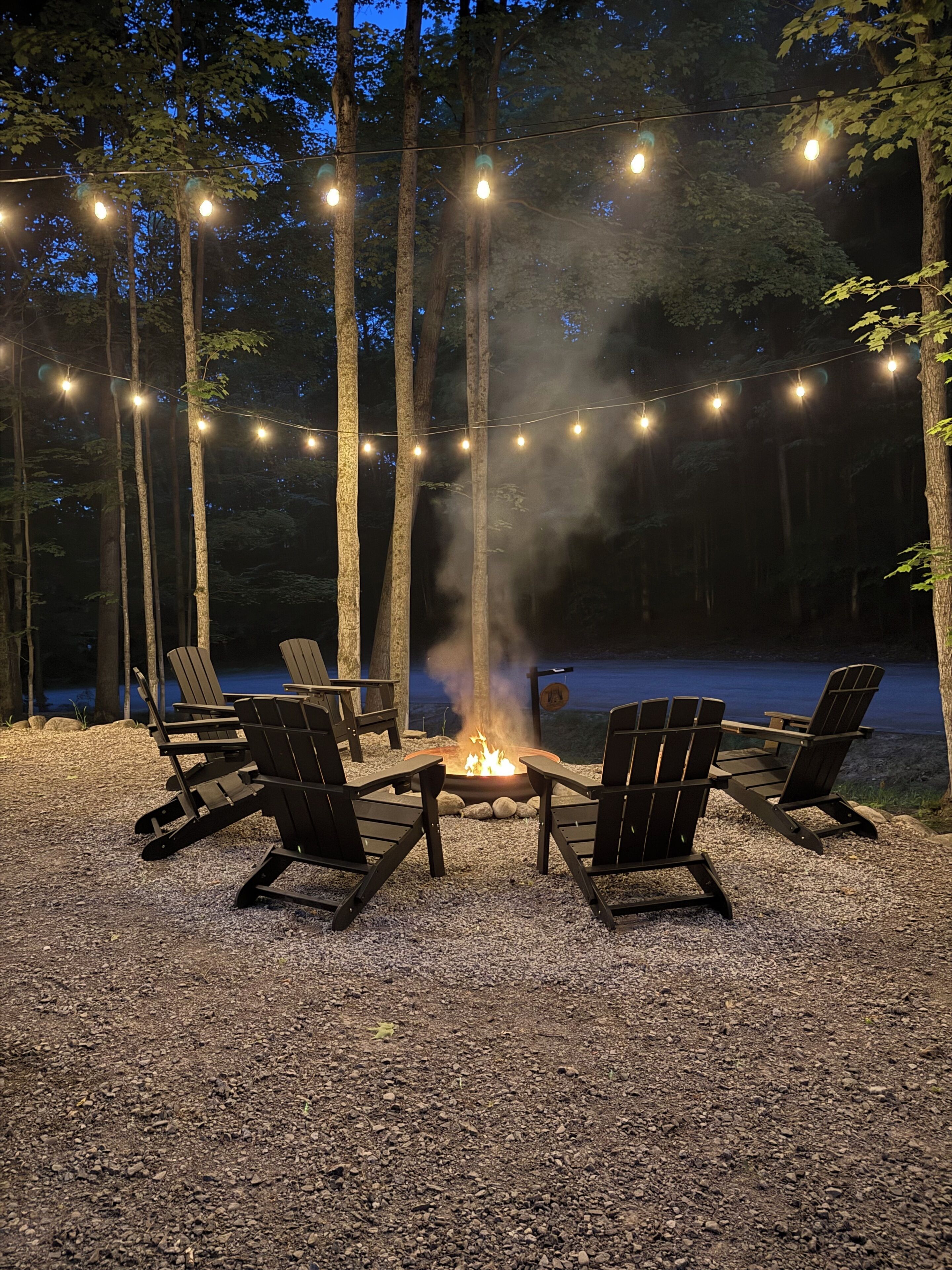 A fire pit with chairs arranged around it, surrounded by trees and string lights.