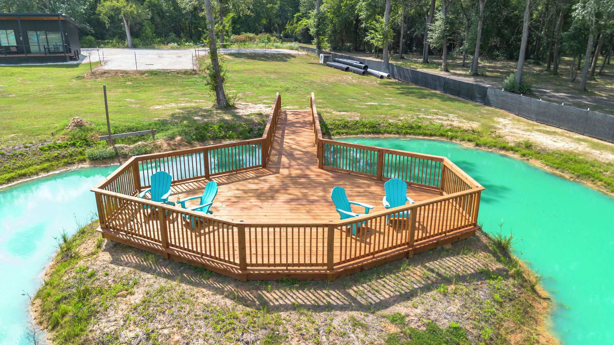 A wooden deck with a view of a turquoise pool, surrounded by a grassy area and trees.