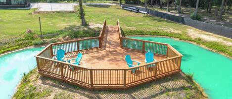 A wooden deck with a view of a turquoise pool, surrounded by a grassy area and trees.