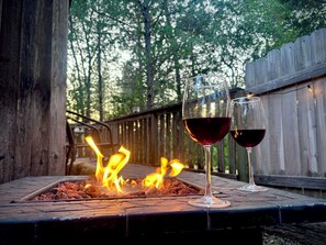 Un foyer extérieur avec deux verres de vin rouge, posés sur une terrasse en bois, entourée d’une clôture en bois et d’arbres.