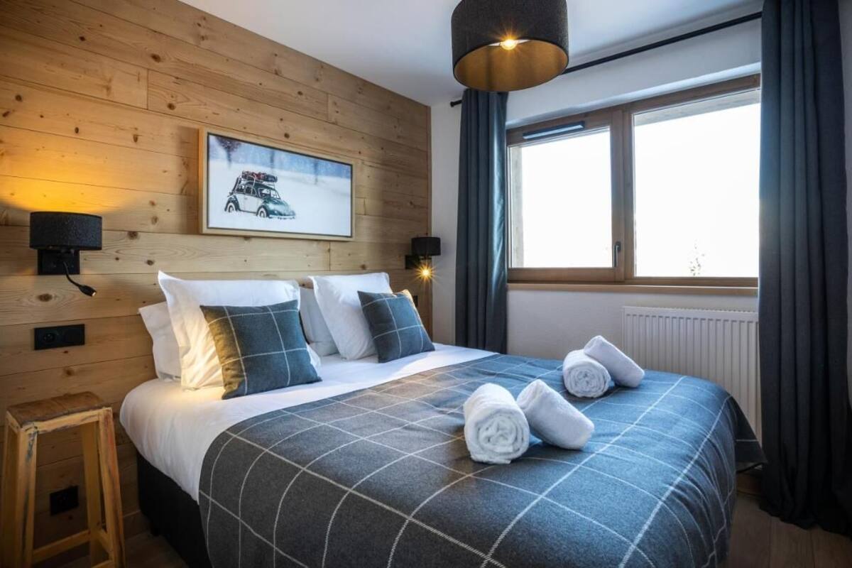 Un lit bien fait, recouvert d’une couvre-lit à carreaux, deux lampes fixées au mur, un tableau encadré accroché au mur et une fenêtre avec des rideaux.
