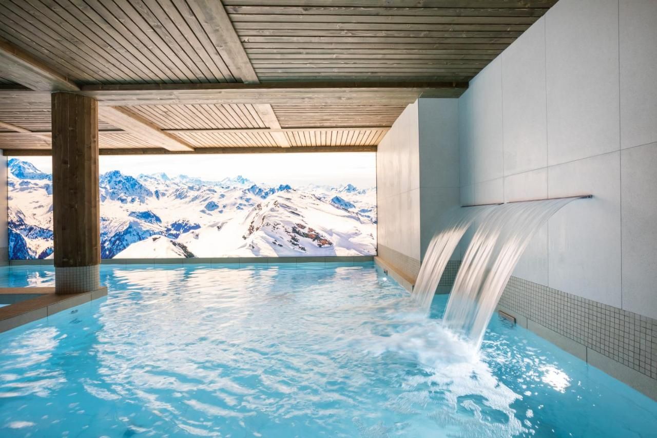 A hot tub with a waterfall feature, surrounded by a wooden ceiling and a view of snow-capped mountains.