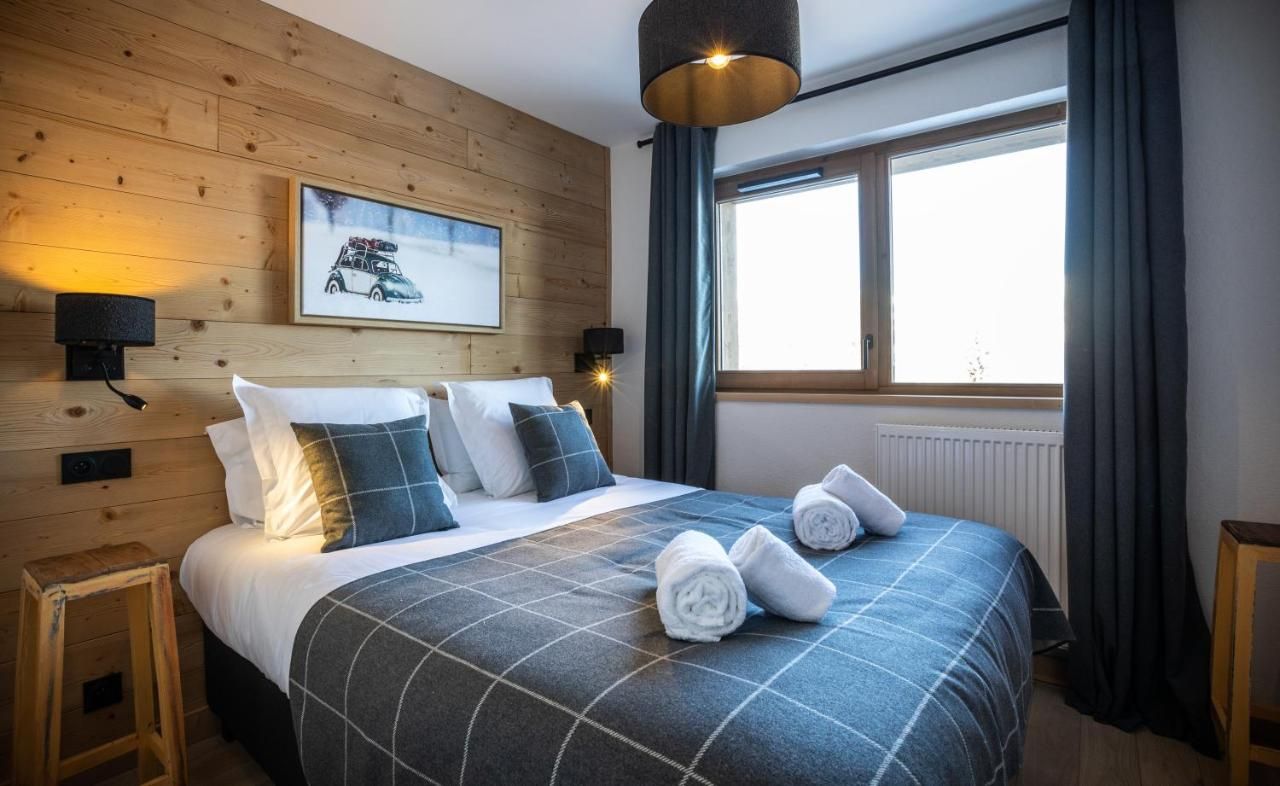 Ein ordentlich bezogenes Bett mit karierter Bettdecke, zwei Wandlampen, ein gerahmtes Bild an der Wand und ein Fenster mit Vorhängen.