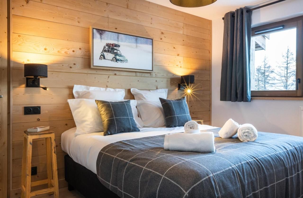 Ein gemütliches Schlafzimmer mit einem Holzkopfteil, einem Bett mit karierter Bettdecke, zwei Wandleuchten, einem gerahmten Bild und einem Fenster mit Vorhängen.