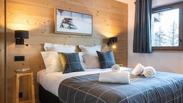 Une chambre confortable avec une tête de lit en bois, un lit recouvert d’une couette à carreaux, deux lampes fixées au mur, un tableau encadré et une fenêtre avec des rideaux.