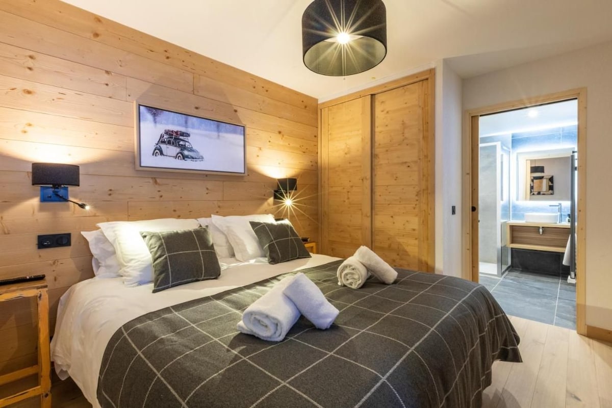 Une chambre d’hôtel avec un mur en bois, un lit recouvert d’une couverture à carreaux, deux oreillers, une serviette pliée, un tableau représentant une voiture accroché au mur, et une table de chevet avec une lampe.