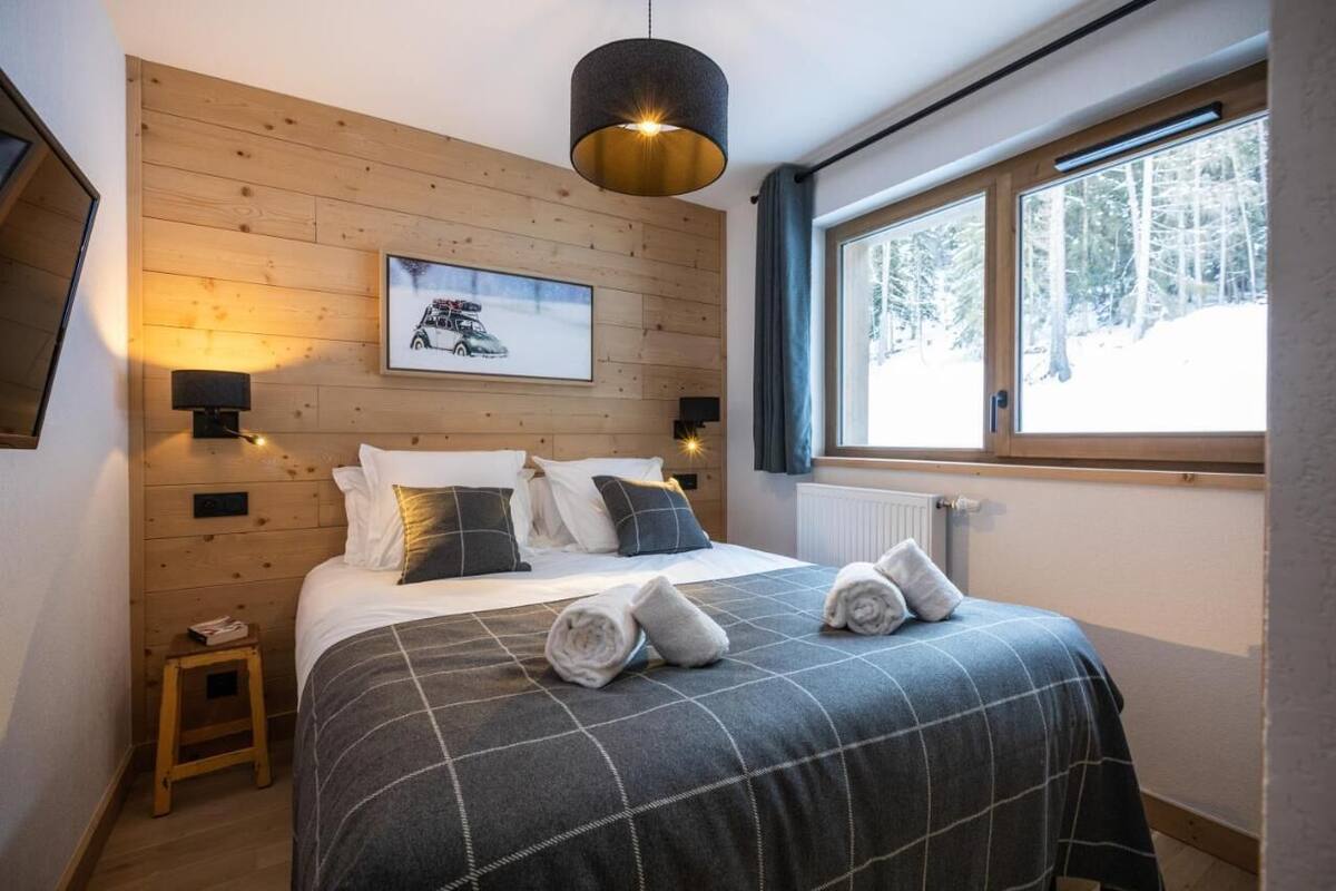 Une chambre avec un mur en bois, un lit recouvert d’une couverture à carreaux, deux lampes de chevet, un tableau encadré au mur et une fenêtre donnant sur des arbres.