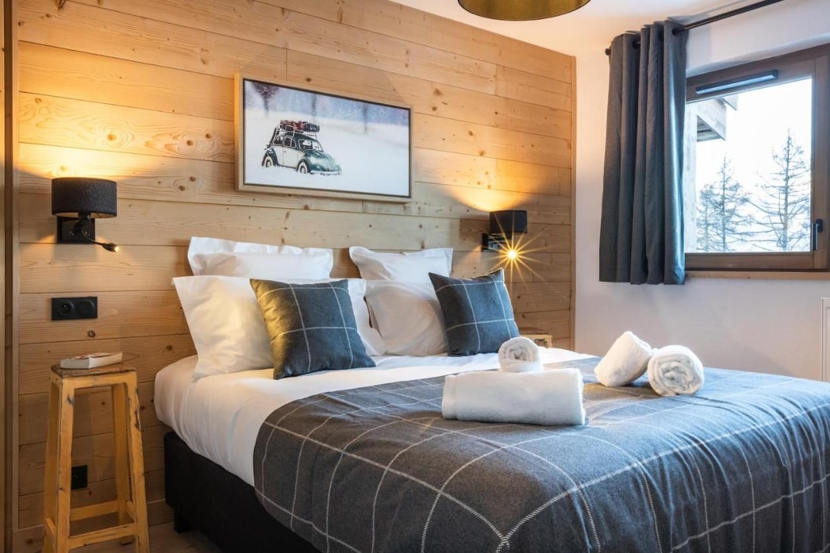 Une chambre confortable avec une tête de lit en bois, un lit recouvert d’une couette à carreaux, deux lampes fixées au mur, un tableau encadré et une fenêtre avec des rideaux.