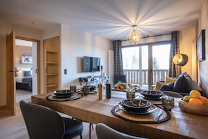 Un espace de repas moderne avec une table en bois dressée pour le repas, un téléviseur fixé au mur et un balcon offrant une vue sur les arbres.