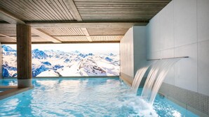 A hot tub with a waterfall feature, surrounded by a wooden ceiling and a view of snow-capped mountains.