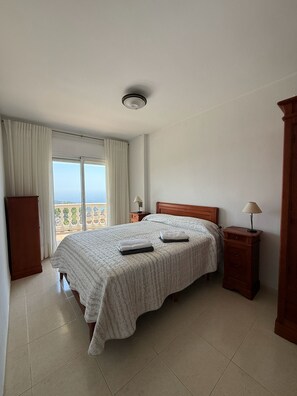 A bedroom with a bed, a nightstand, a lamp, a wardrobe, and a view of the sea through a window.