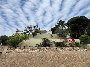 A stone wall with a garden in front, a building with a sloped roof, and trees in the background.