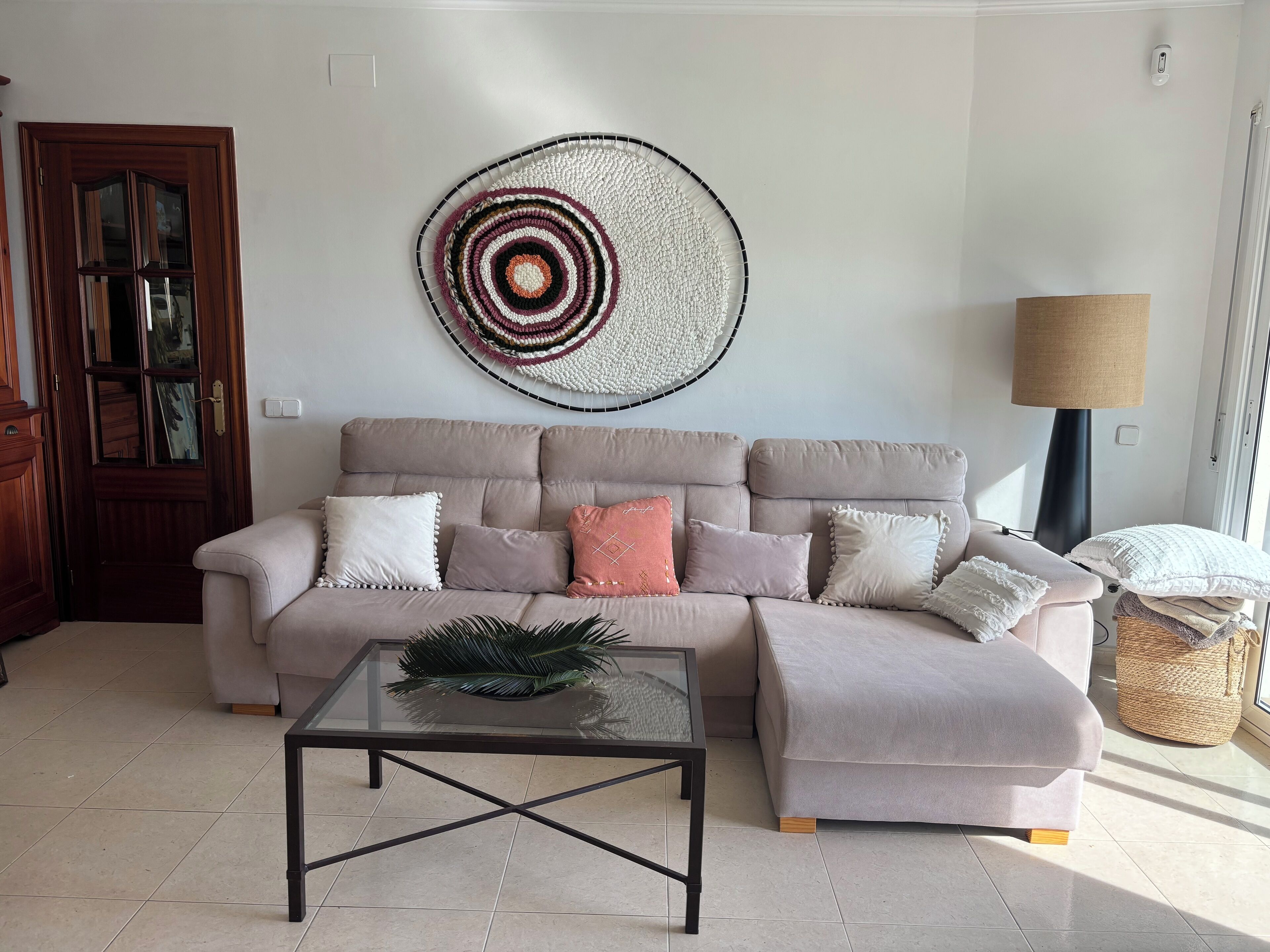 A living room with a beige sectional sofa, a glass coffee table, and a wall-mounted circular woven artwork.