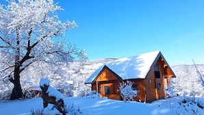 A wooden cabin with a snow-covered roof, surrounded by snow-covered trees and a clear blue sky.